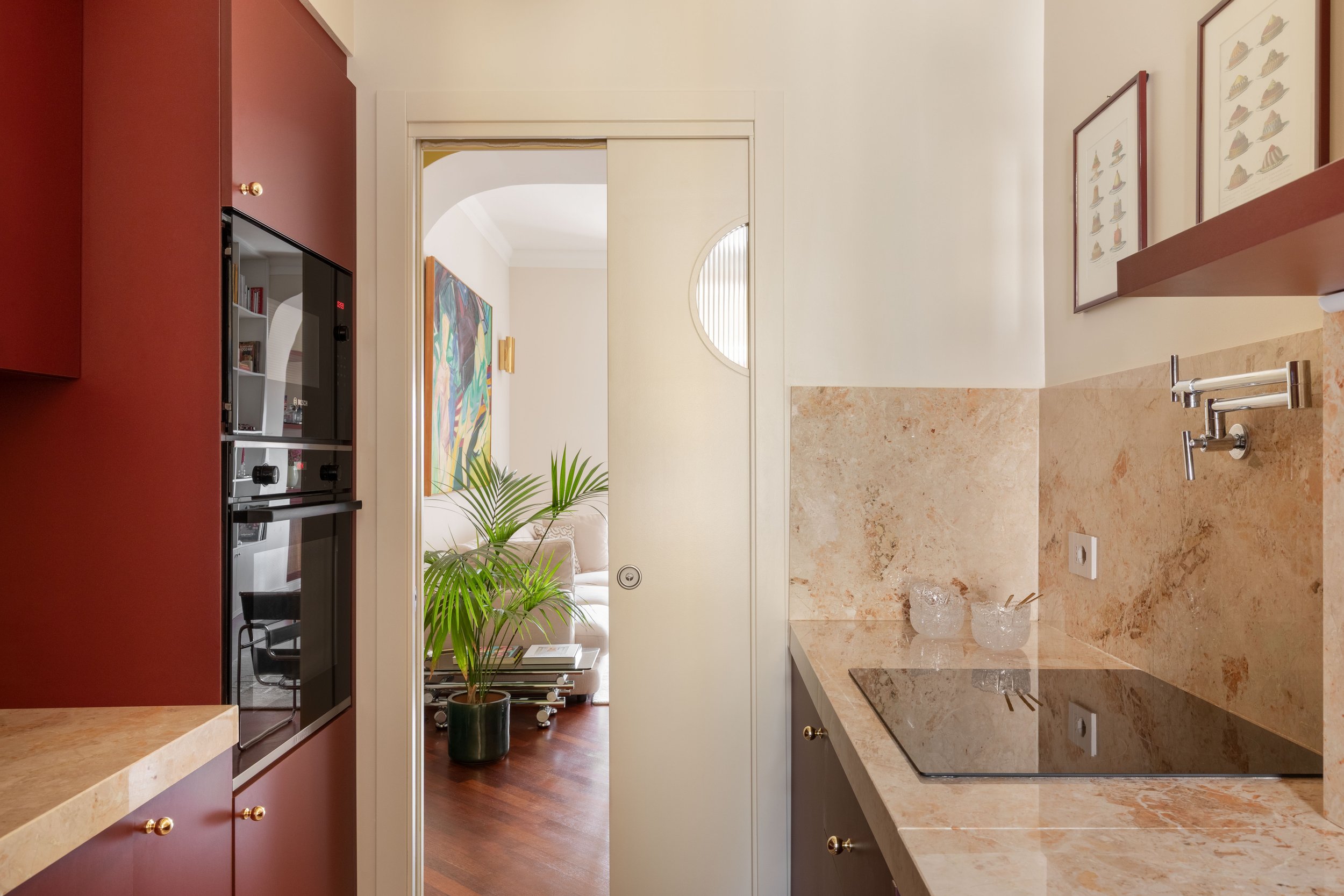 Interior di una cucina con ante rosse, pareti chiare, piano di lavoro in marmo, fregi e quadri alle pareti, e un'area che porta a un soggiorno con un dipinto colorato e una pianta verde.