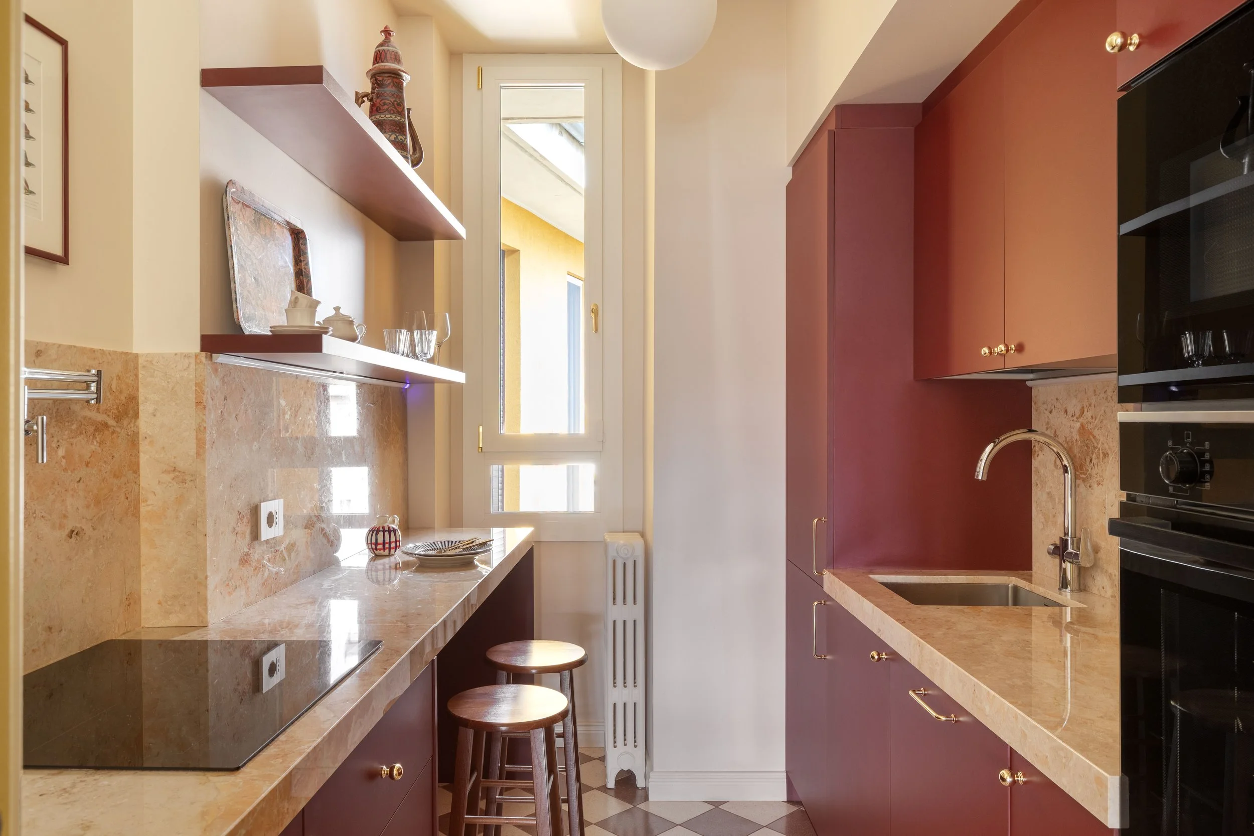 Una cucina con ante rosse, un bancone in marmo e un piano cottura nero. La finestra lascia entrare luce naturale e ci sono sgabelli di legno ai lati del bancone.