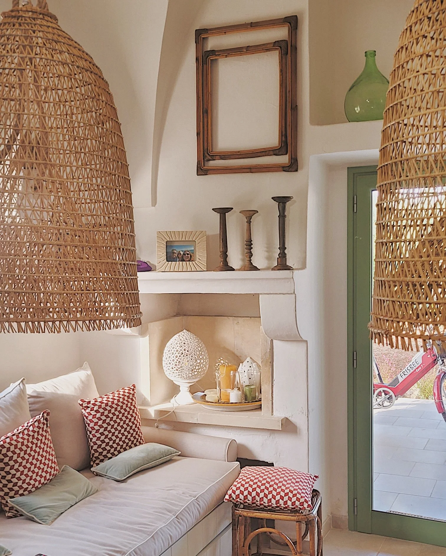 Interno soggiorno con divano bianco, cuscini a motivo geometrico rosso e bianco, lampada ornamentale sopra il camino, mensole con candeliere, cornice con foto, porta finestra verde parzialmente aperta.