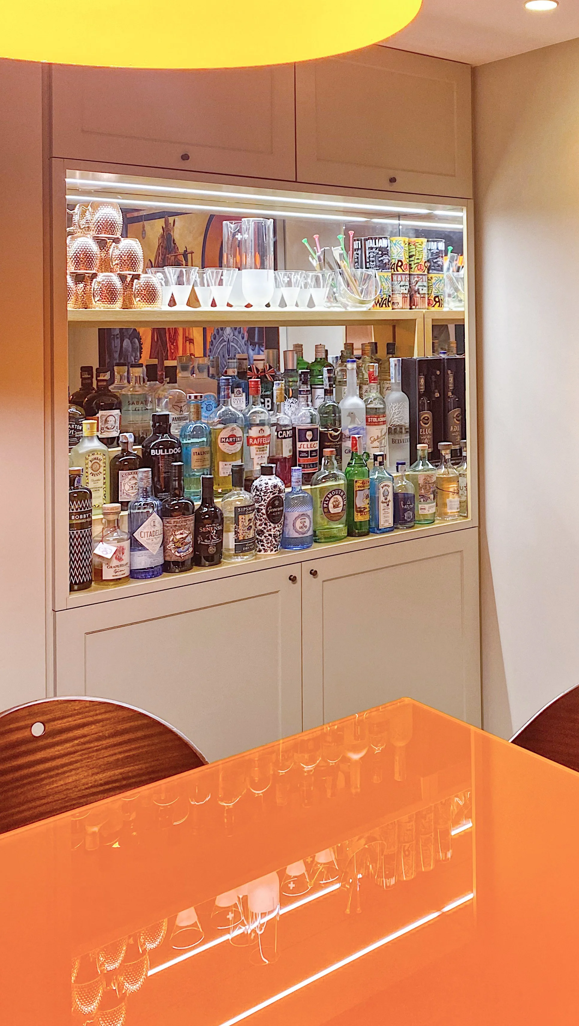 Uno scaffale con bottiglie di alcolici, bicchieri e accessori per cocktail, in un bar o ristorante, con luci soffuse ed un tavolo di colore arancione in primo piano.