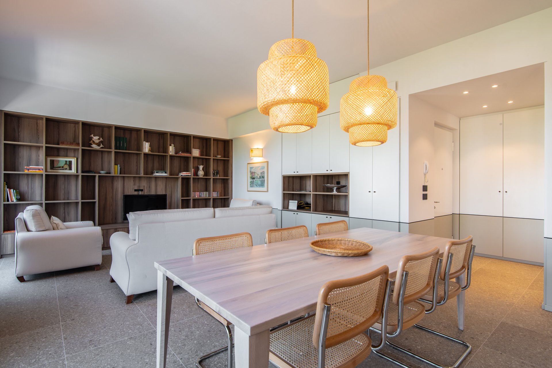 Soggiorno con tavolo e sedie in primo piano, divani bianchi e libreria in legno scuro sullo sfondo, lampade di vimini sospese sopra il tavolo, pareti biene con quadri e scaffali.