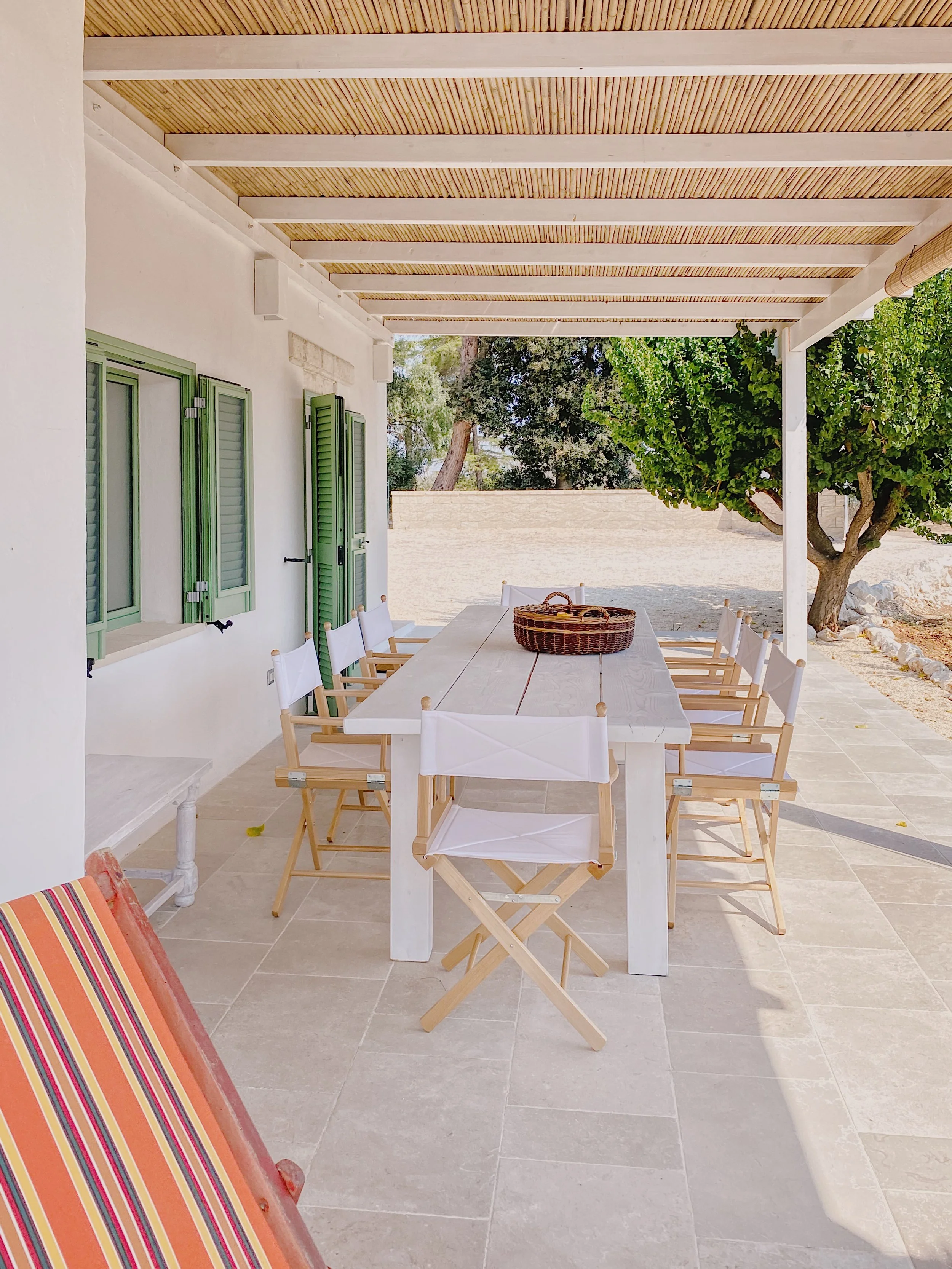 Veranda estiva con tavolo bianco e sedie, coperta con pali di bambù, vicino a una casa bianca con persiane verdi, con uccello di vimini sul tavolo e una sedia pieghevole con sedie di legno chiaro, circondata da alberi e arbusti.