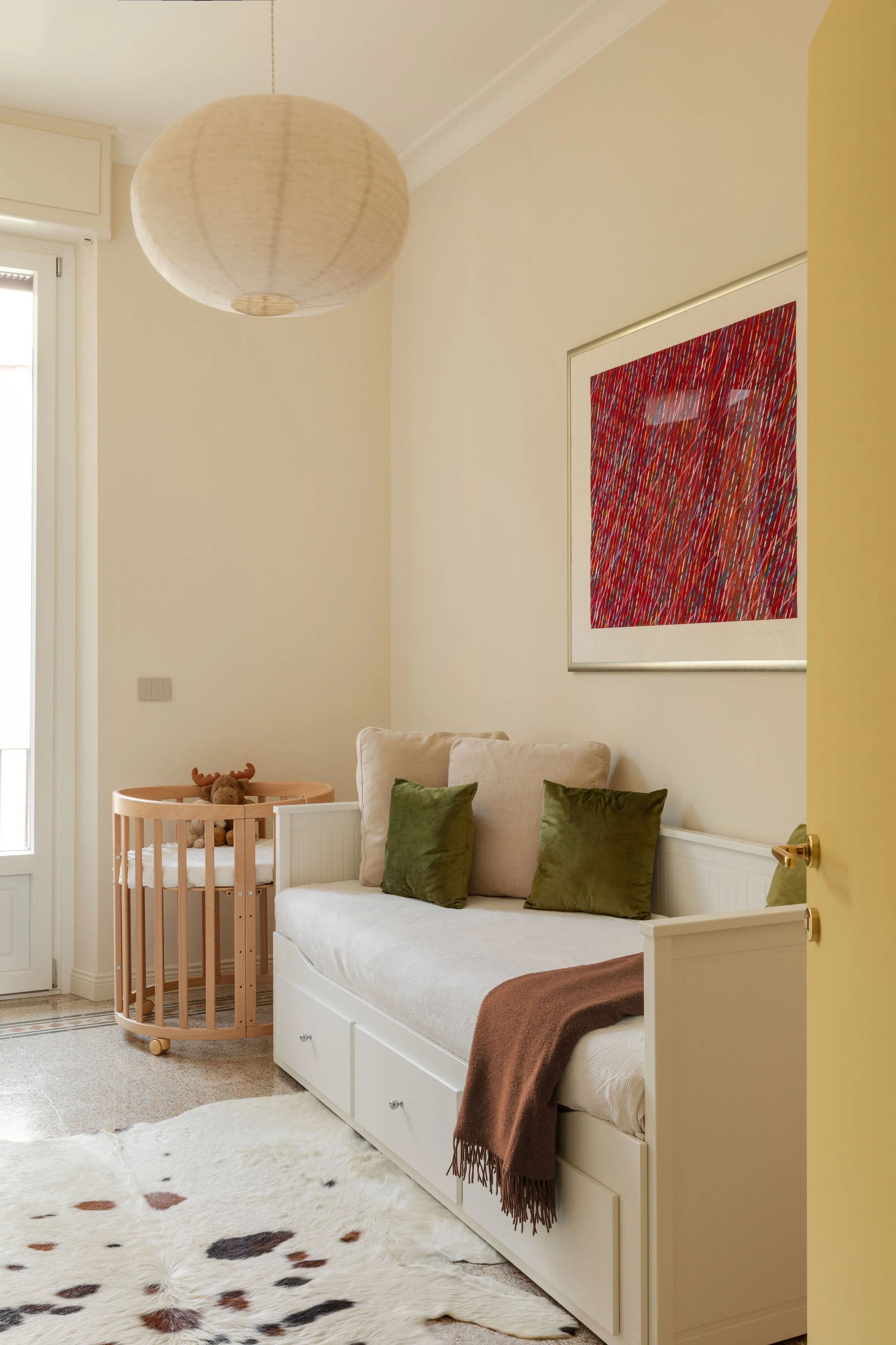 Angolo soggiorno con divano bianco, cuscini verdi, stampa astratta rossa e beige, lampada a soffitto rotonda, ingresso naturale, tappeto con macchie nere, adiacente lettino per bambini con giocattolo orso. Pareti di colore crema e giallo chiaro.