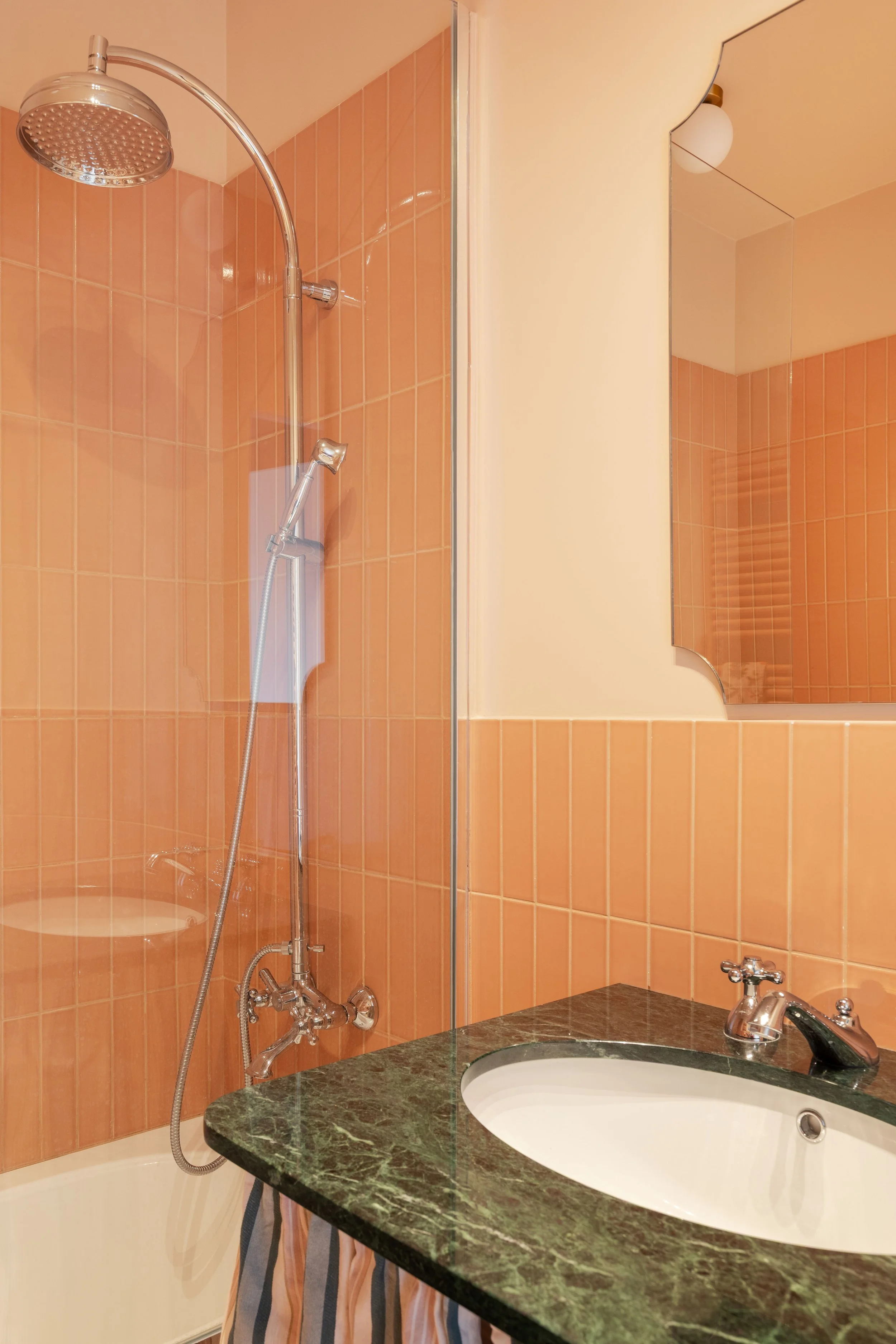 Bagno con piastrelle arancioni, lavandino con top in marmo verde scuro, specchio sopra il lavandino, doccia con soffione e parete divisoria di vetro, lavabo e rubinetto, lampada bianca a soffitto.