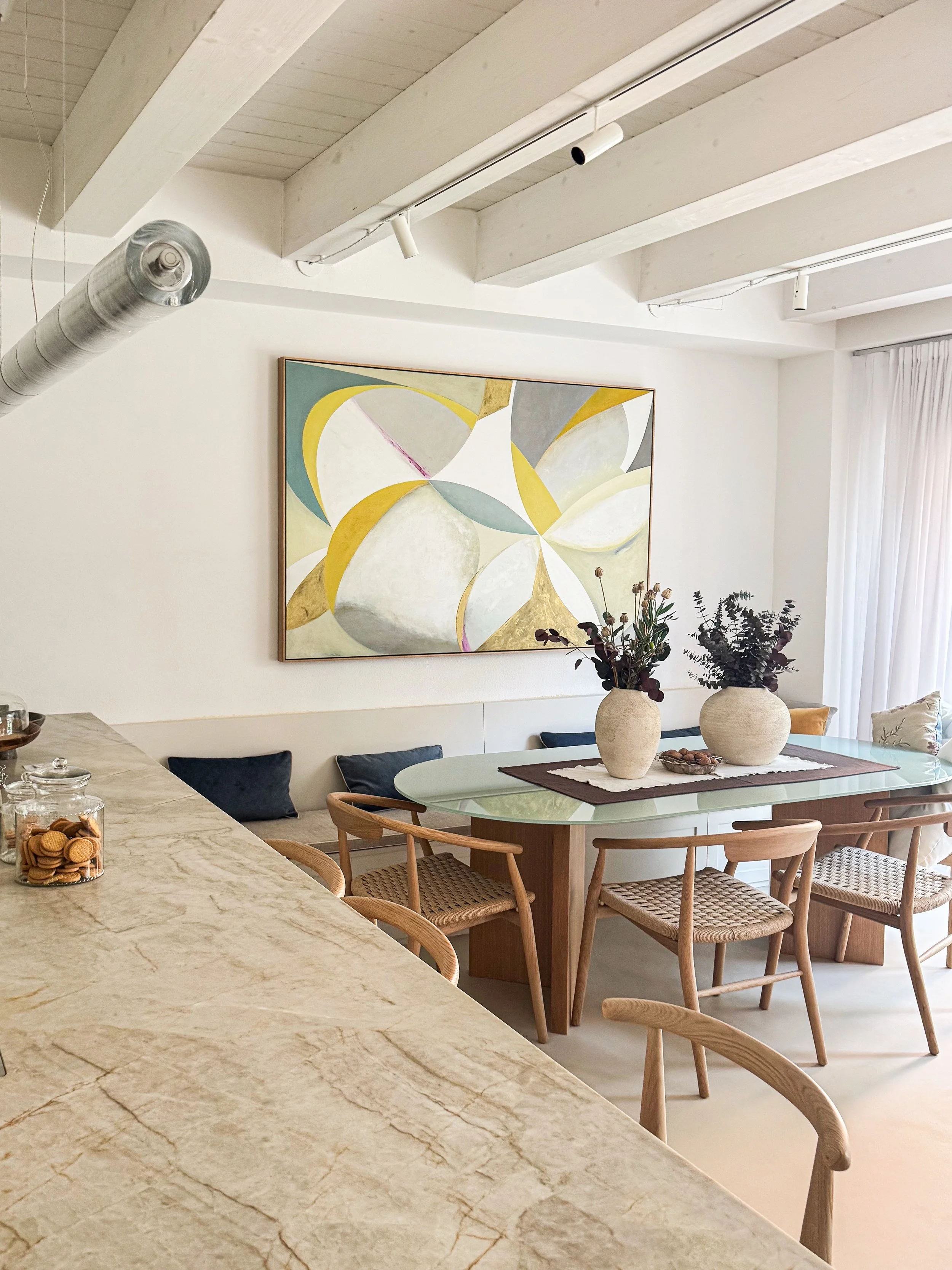 Interno di una sala da pranzo moderna con un grande dipinto astratto sulle pareti, un tavolo rotondo con vasi di fiori e sedie in legno con sedute in vimini.