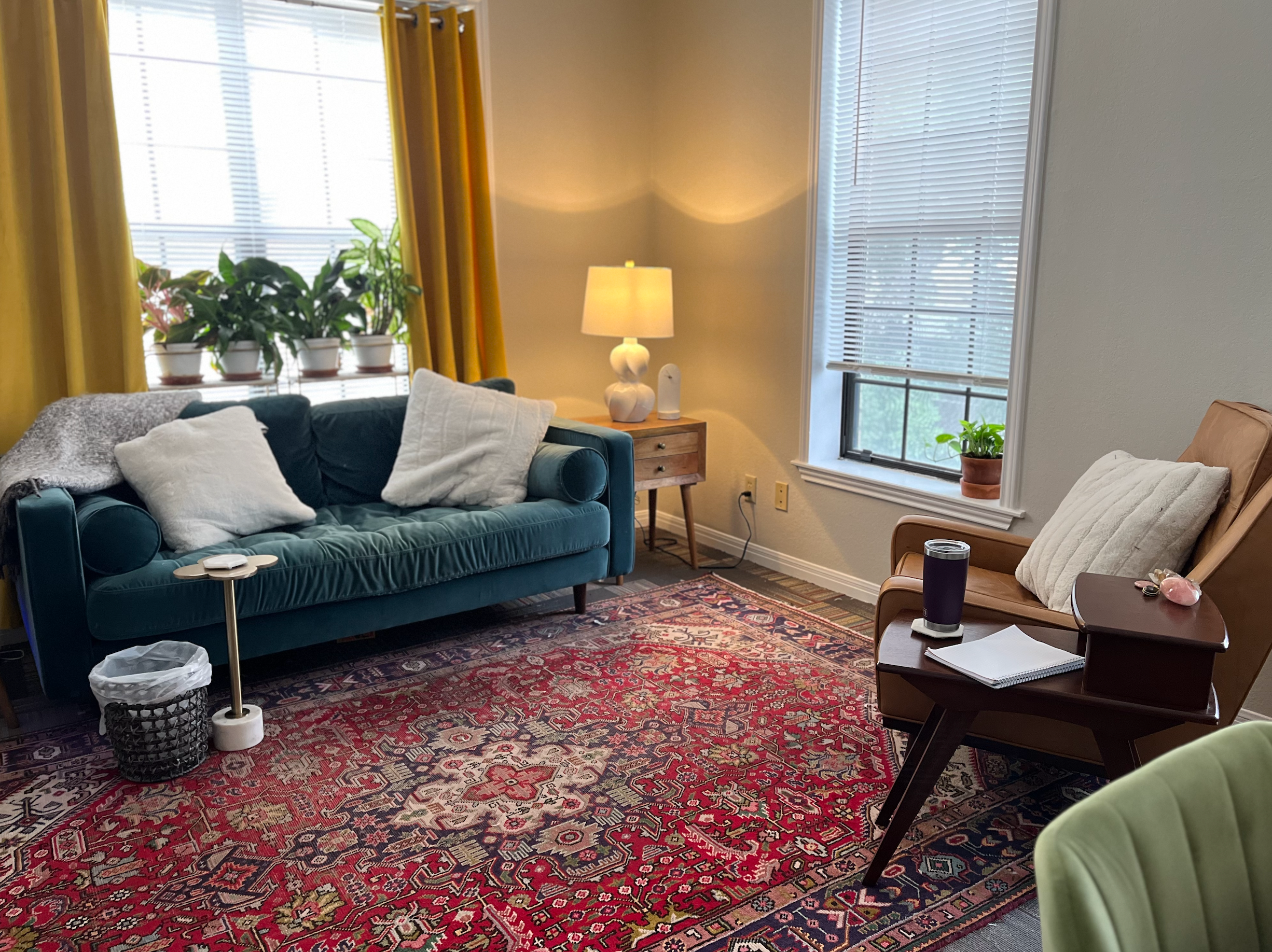 A cozy living room with a teal sofa, cream pillows, and a gray throw blanket, a wooden side table with a white lamp, a patterned area rug, and a brown armchair with a pillow. The room has two large windows with blinds, yellow curtains on one, and potted plants.