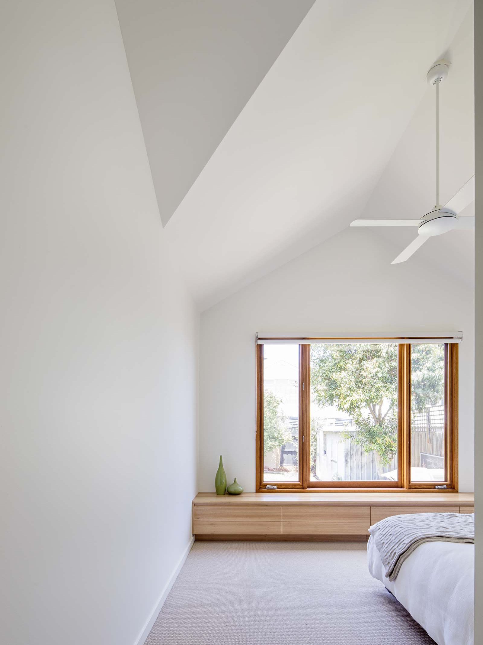 A minimal bedroom with white walls, a large window with wooden frame showing greenery outside, a wooden shelf with three decorative vases, and part of a bed with a light-colored bedspread.