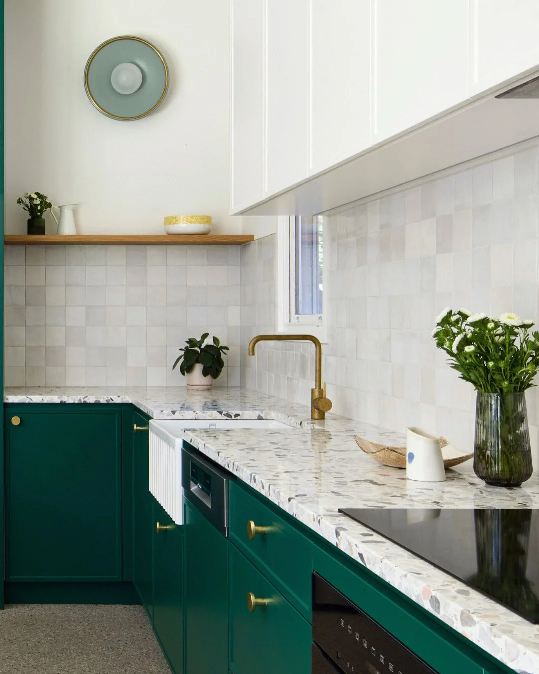 Modern kitchen with green cabinets, white marble countertop, gold faucet, potted plants, and decorative items on shelves and counters.