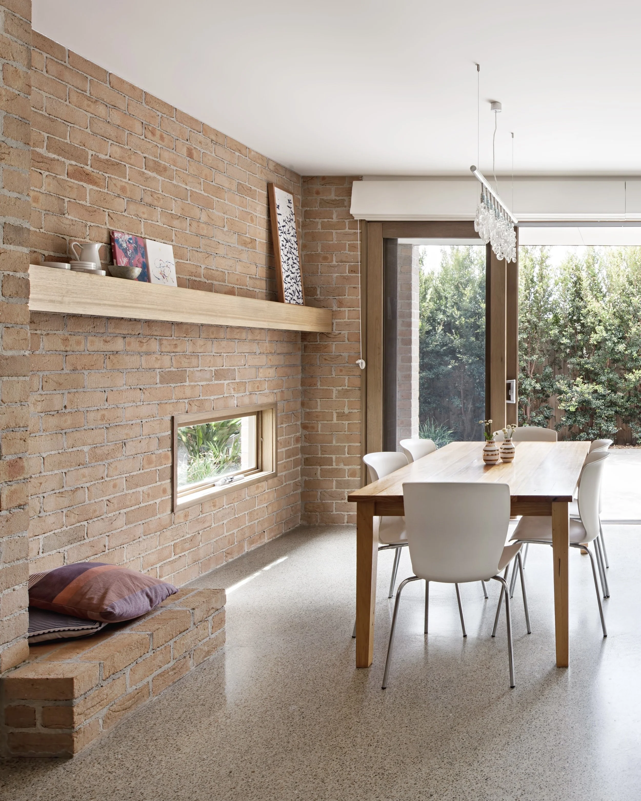 A dining room with a brick wall, a wooden table, white chairs, and a large window showing outdoor greenery. Decor includes vases on the table and artwork on the brick wall, with a crystal chandelier hanging from the ceiling.