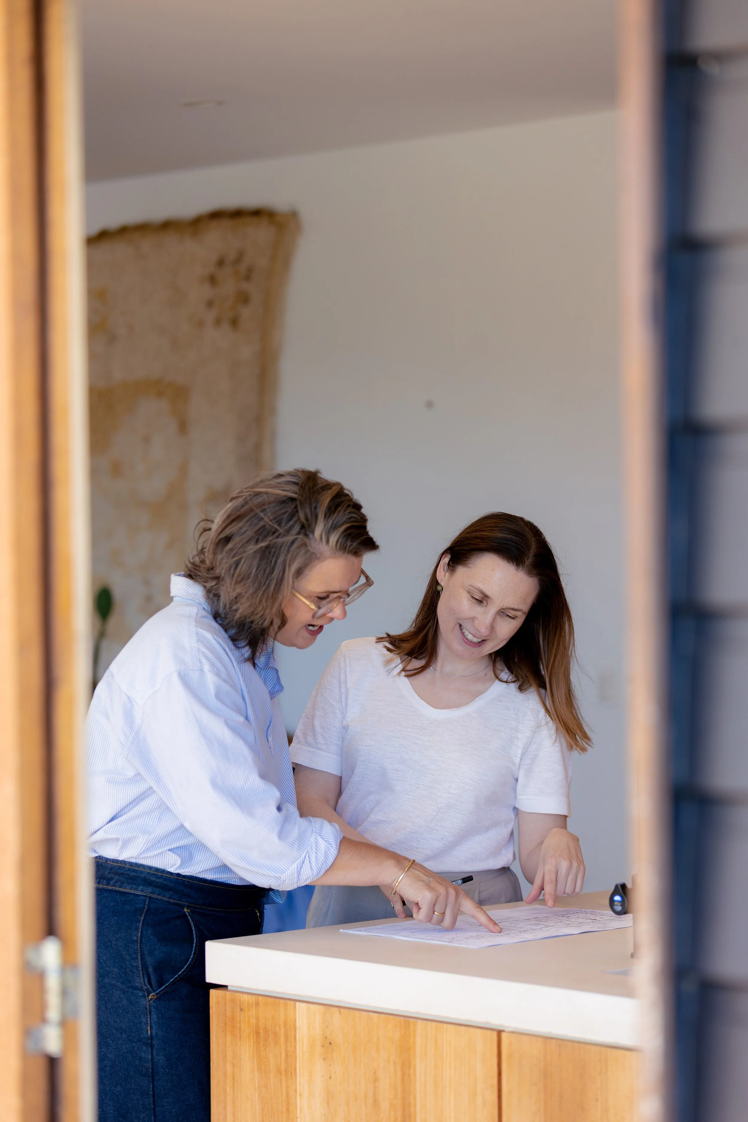 Two women are standing and pointing at a sheet of paper on a white table, engaged in discussion. The photo is taken through a doorway, with the doorway frame visible on the left and right sides.