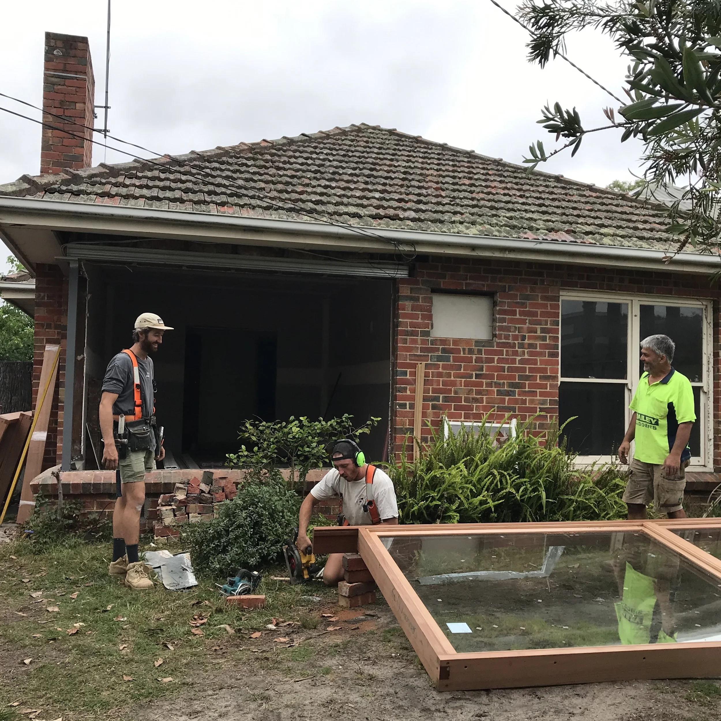 Two men working on a house renovation outside, one in a neon green shirt and another with a beard and hat, surrounded by tools and building materials, with a brick house and a roof in the background.