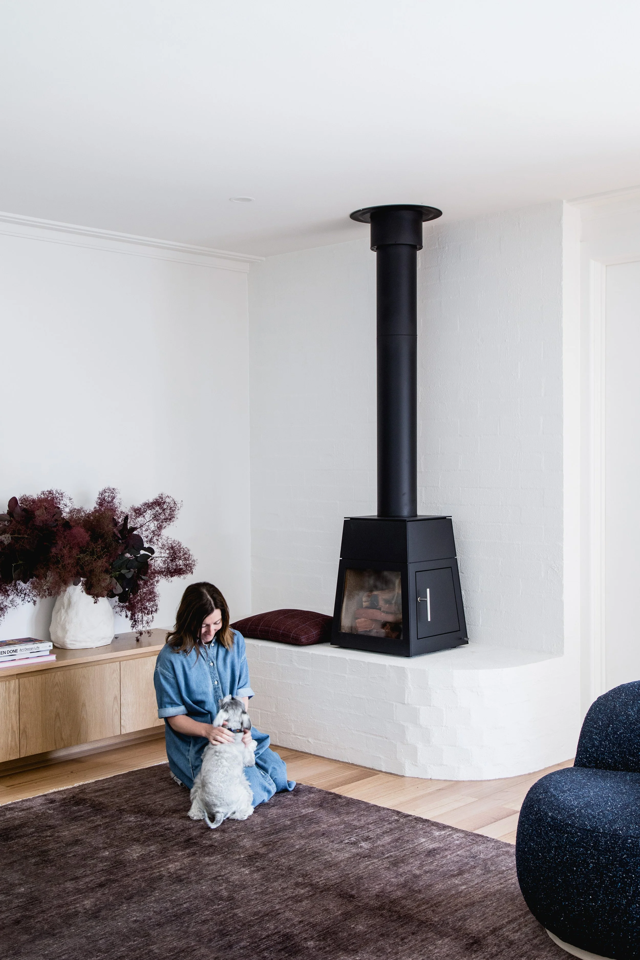 A woman sitting on a brown rug in a cozy living room, playing with a small dog. The room has a white wall, a black wood-burning stove with a tall pipe, a wooden sideboard with a large bouquet of dark red flowers, and a few books. There is a pillow on