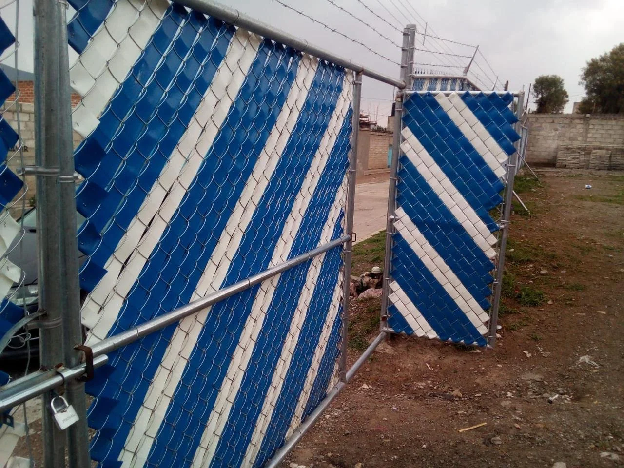 Puerta de malla ciclónica con franjas azules y blancas, cerrada con un candado, en un entorno exterior.