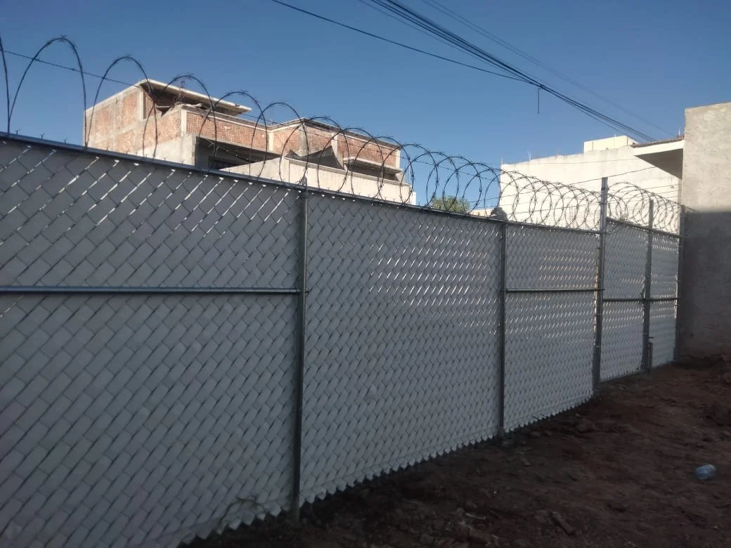 Cerca de alambre cubierto con tela metálica y alambre de púas en la parte superior, frente a edificios de concreto bajo un cielo azul.
