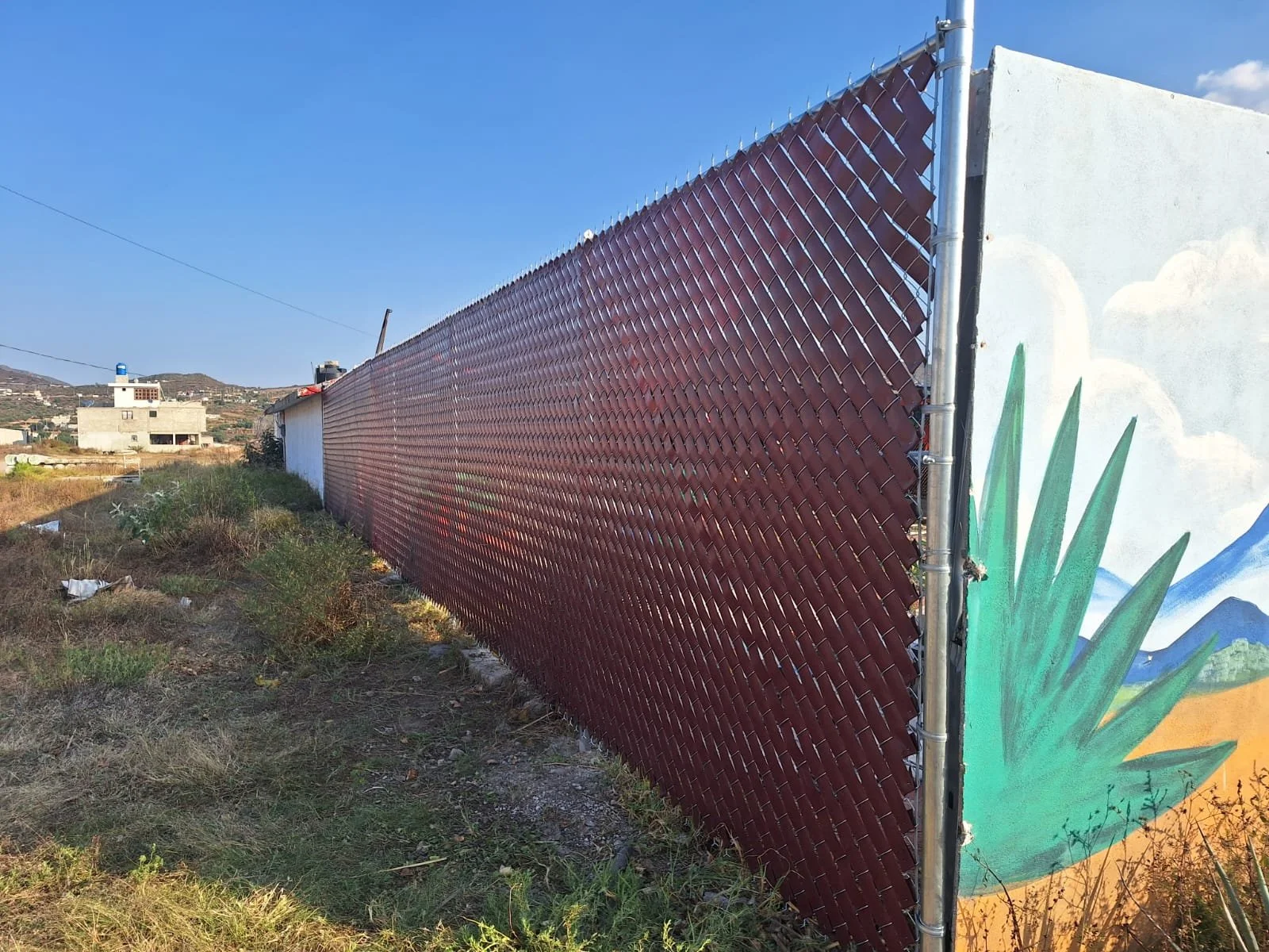 Valla de metal oxidado con pintura en mural artístico en un fondo de paisaje rural, con casas en el horizonte y cielo despejado.