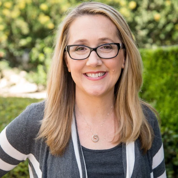 Smiling person with glasses and long blonde hair, wearing a striped cardigan and necklace, outdoors with greenery in the background.