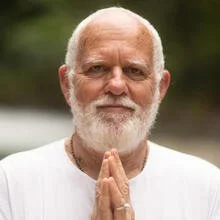 Elderly man with white beard and white shirt, hands together in a gesture of prayer or greeting, outdoors.