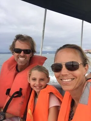Three smiling people, two women and a girl, wearing orange life jackets and sunglasses on a boat with an overcast sky and water in the background.