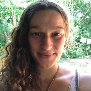 A young woman with long, wavy brown hair smiling outdoors with greenery in the background.