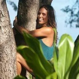 A woman smiling and hugging a tree outdoors surrounded by greenery.