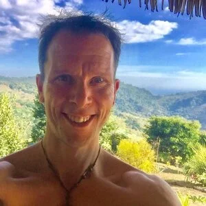 Smiling man with short dark hair taking a selfie outdoors with a lush green landscape and hills in the background under a blue sky with some clouds.