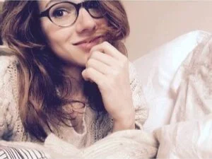 A woman with long brown hair and glasses smiling while resting her chin on her hand, sitting on a bed with light-colored bedding.