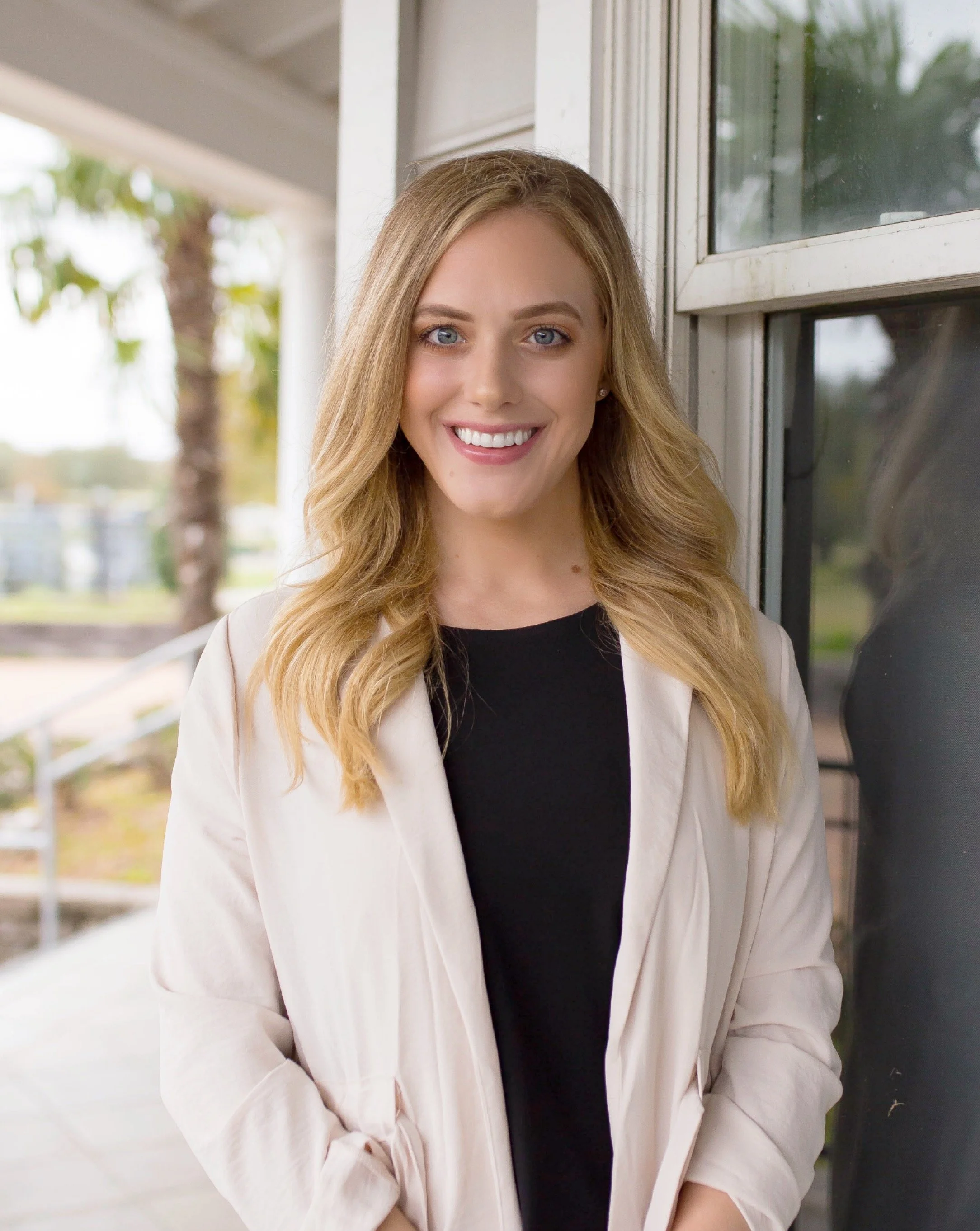Young woman with blonde hair smiling, standing outside near a window.
