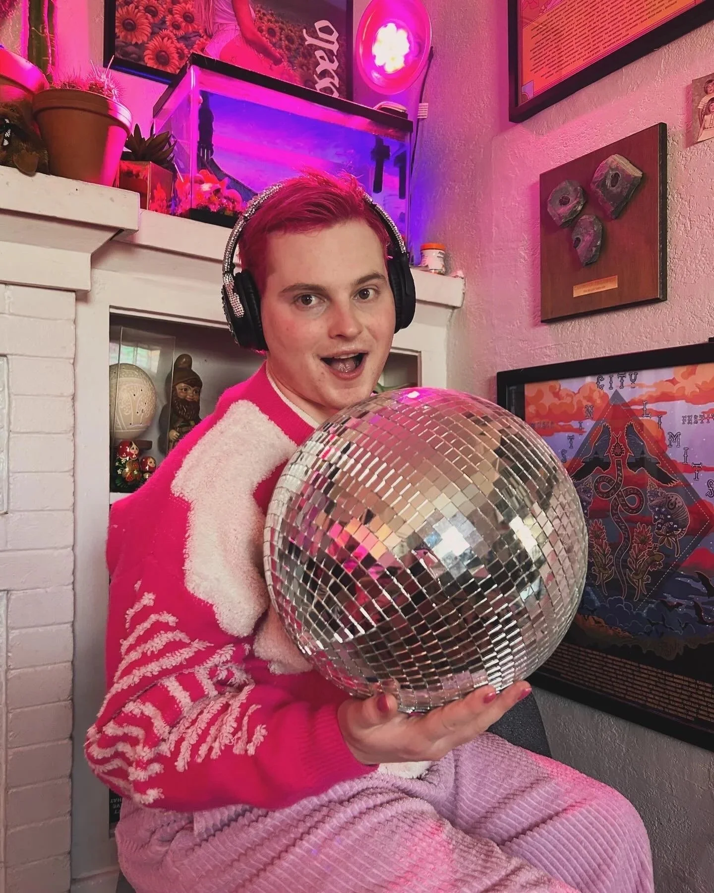 A young person with pink hair and headphones holding a disco ball in a room decorated with pink and purple lighting, plants, and framed artwork.