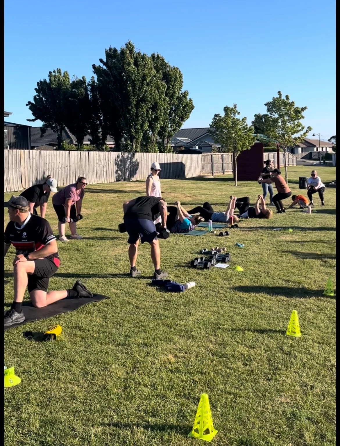 Outdoor fitness class with people exercising on mats, using weights and resistance bands, surrounded by green grass and trees, sky clear.
