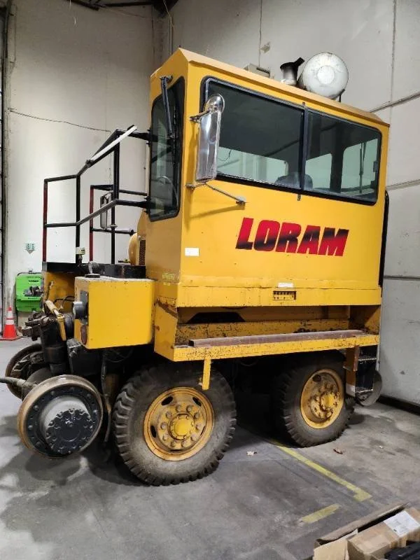 Yellow Loram rail vehicle with a large enclosed operator cabin, large tires, and mechanical parts in an indoor storage area.
