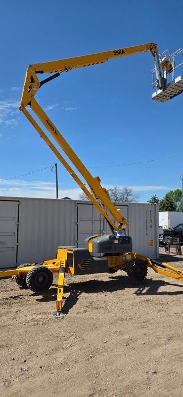 A yellow telescopic boom lift with extended arm and platform at the top, positioned on a dirt ground outdoors.