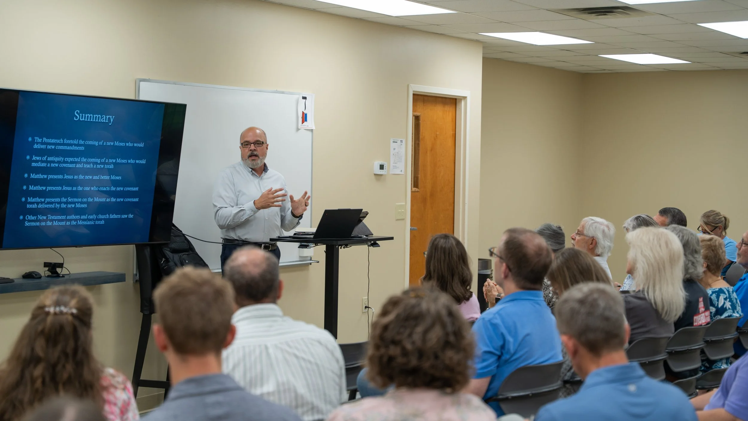 A man standing in front of a whiteboard giving a presentation in a classroom or seminar room, with an audience seated and listening.