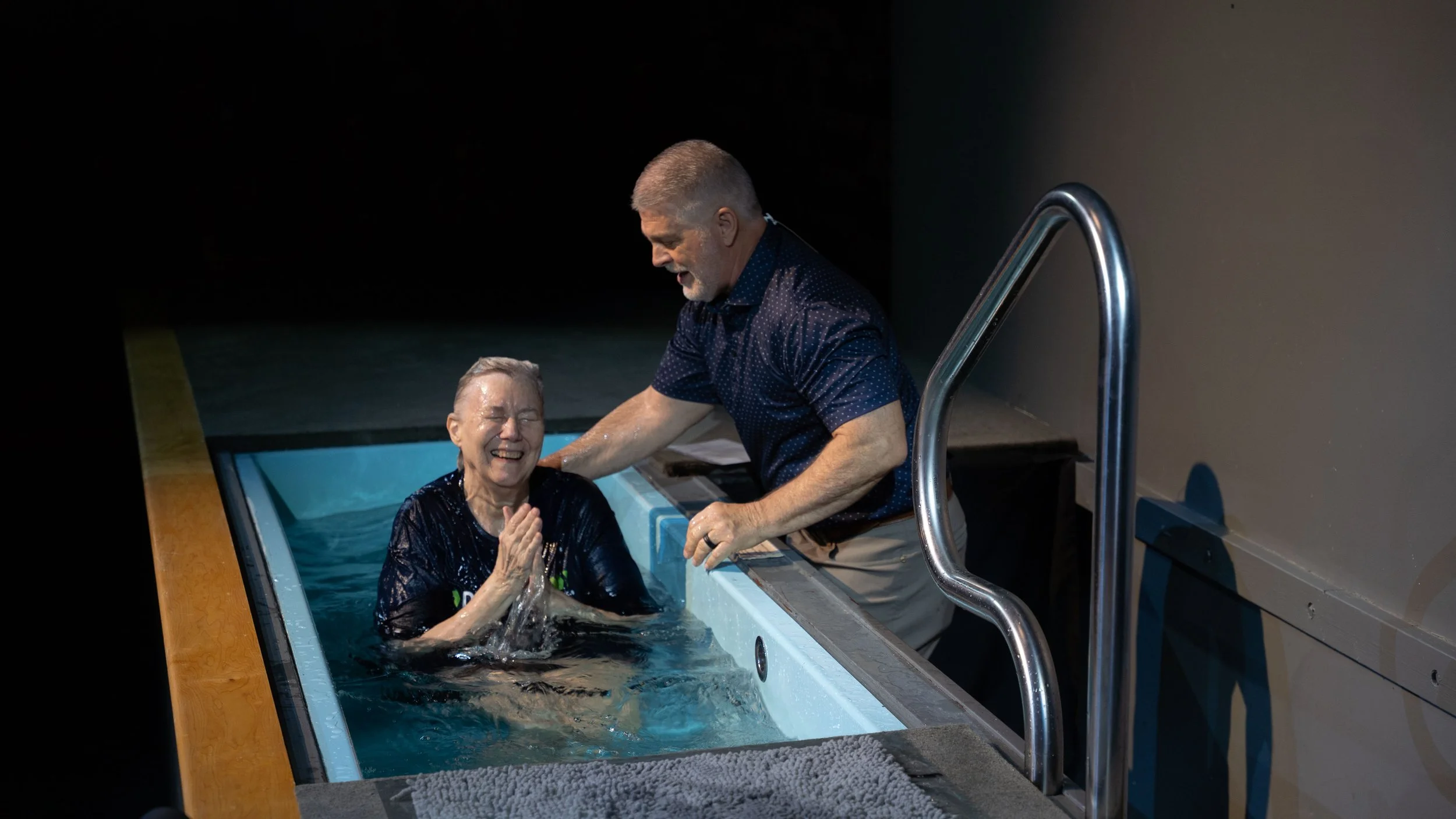 A woman in a baptismal pool with her arms raised, smiling, wearing a blue shirt that says 'DISCIPLE'. A man stands beside her, smiling, wearing glasses, a striped shirt, and a blue vest. The background is dimly lit with a blue wall and a ladder leading into the pool.