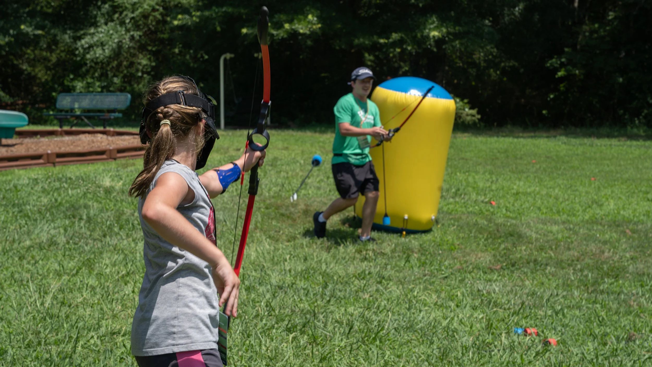 A young boy participates in a bubble archery activity outdoors on a grassy field, aiming a toy bow at targets, with inflatable barriers and trees in the background.