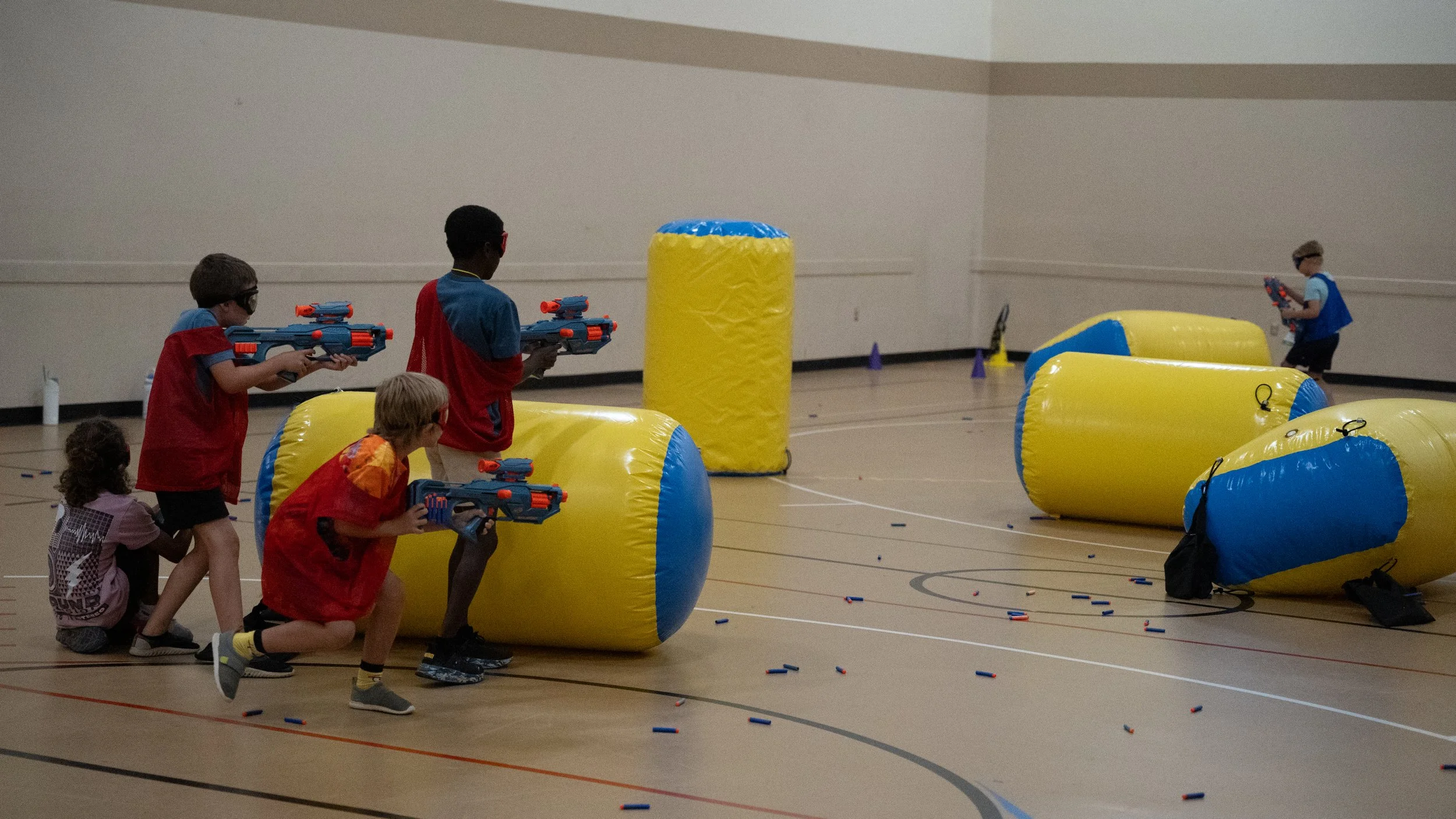 Children wearing protective goggles playing nerf gun game in a gymnasium with inflatable barriers.