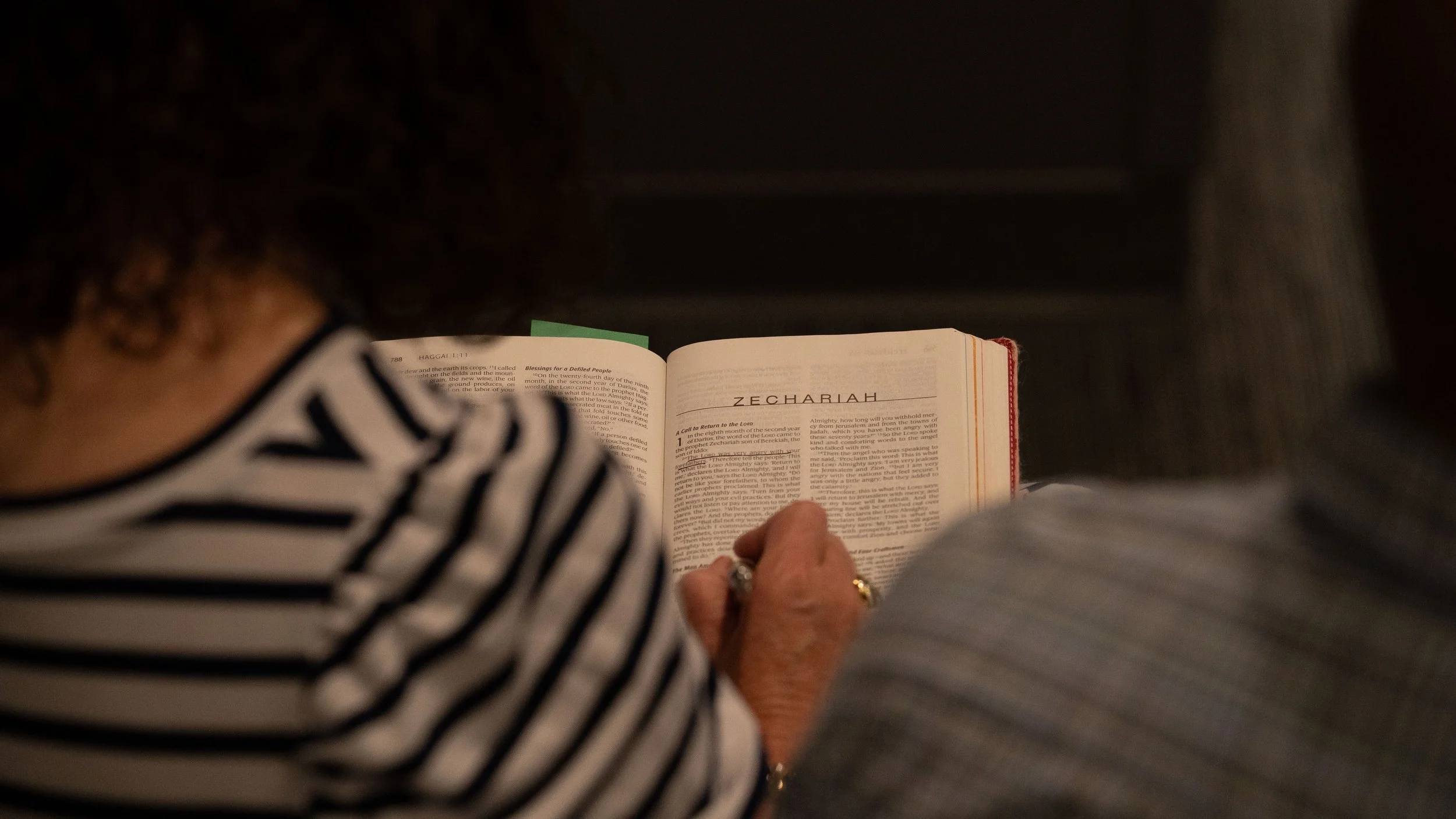 Person sitting in a chair reading a Bible, wearing a green striped shirt, blue pants, brown shoes, and a watch, in a setting that appears to be a church or auditorium.