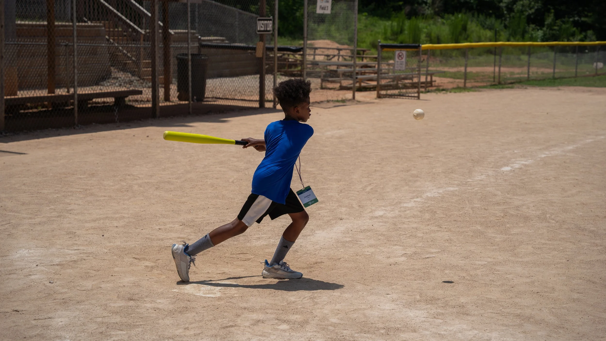 A young girl in an orange shirt and white sneakers is playing wiffleball on a dirt field, holding a blue bat, preparing to hit a white ball flying through the air, with a chain-link fence and bleachers in the background.