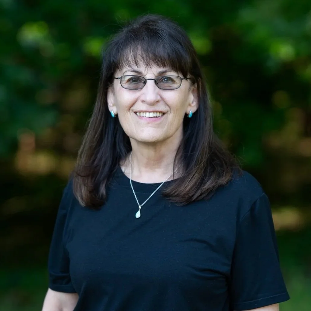 A smiling woman with black hair, glasses, and hoop earrings, wearing a pink sleeveless top, standing outdoors with green trees and grass in the background.