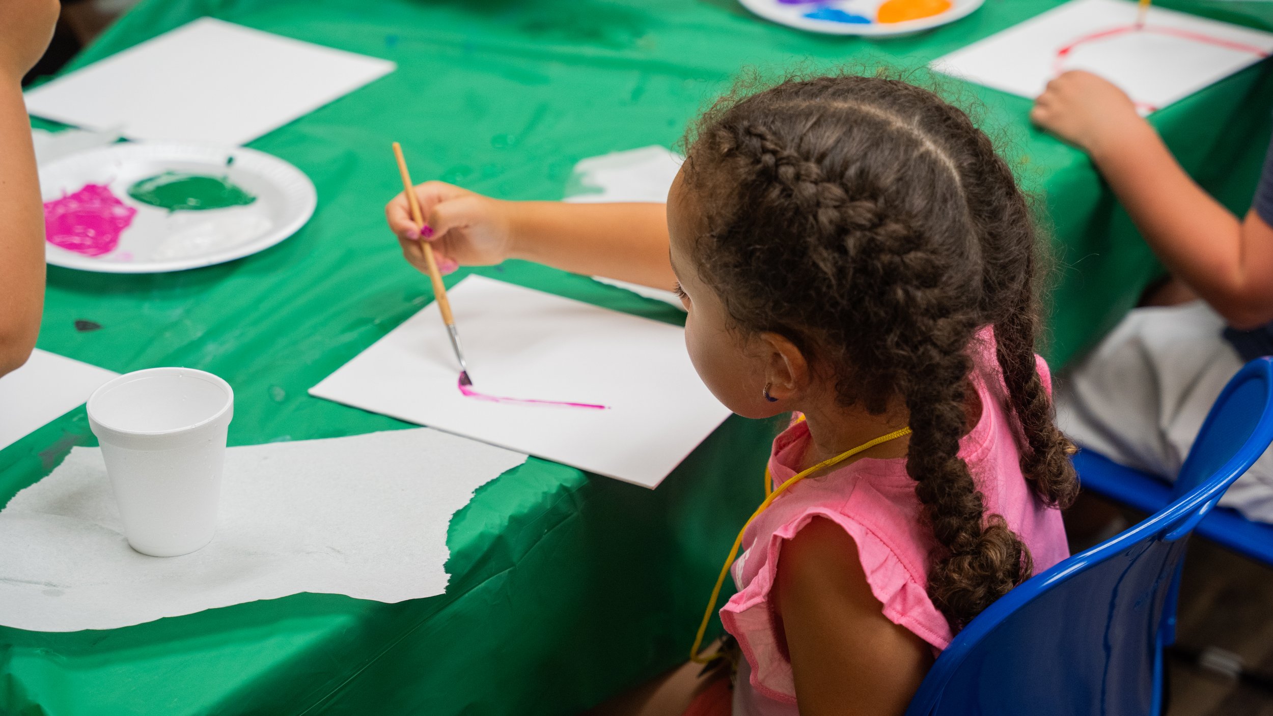 Children painting pictures at a table during an art activity, wearing white smocks with yellow tablecloths and various art supplies.