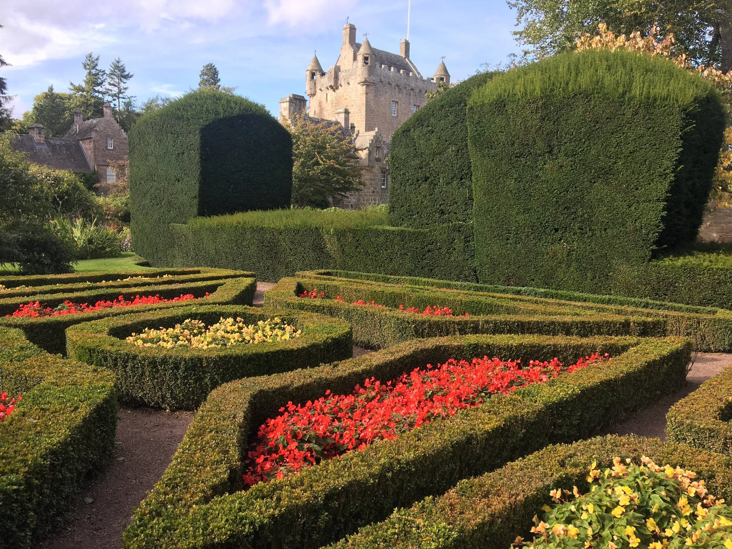 Beautiful garden with neatly trimmed hedges and colorful flowers in front of an old stone building, possibly a castle, surrounded by trees.