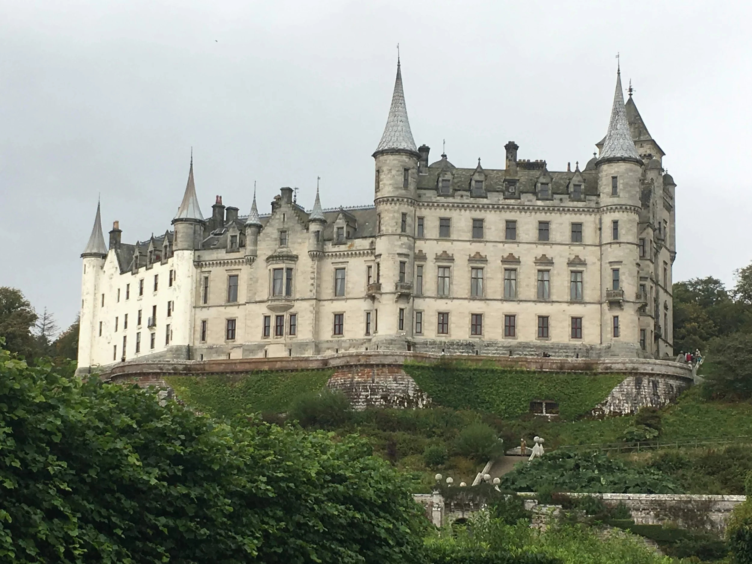 Historic castle with numerous towers and turrets, surrounded by greenery, under an overcast sky.