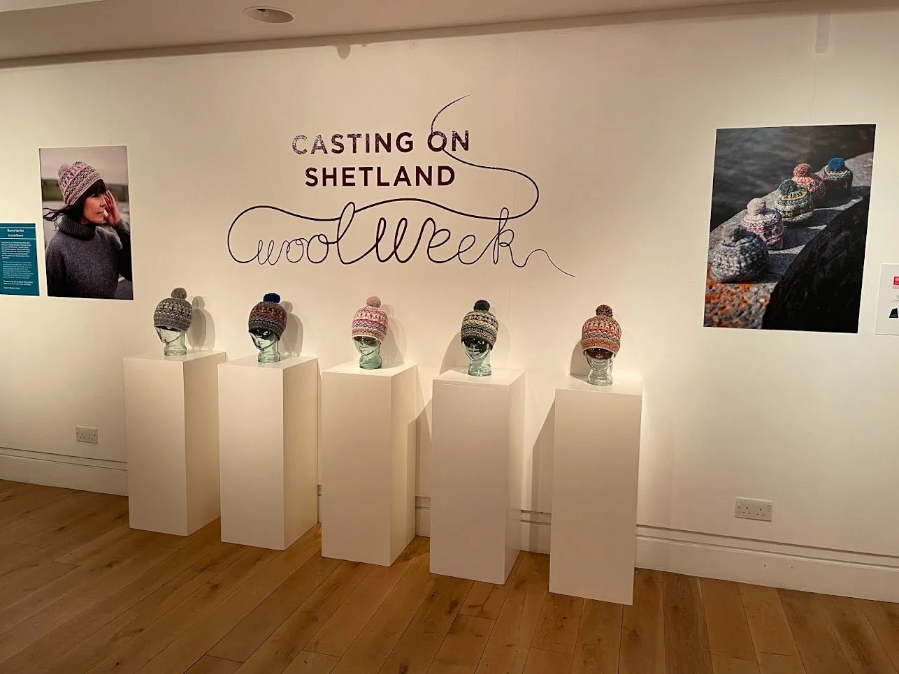 Exhibition display featuring knitted hats on white pedestals, wall text reading "Casting on Shetland Wool Week," and photos of hats and a person wearing one. Wood floor and plain white walls.