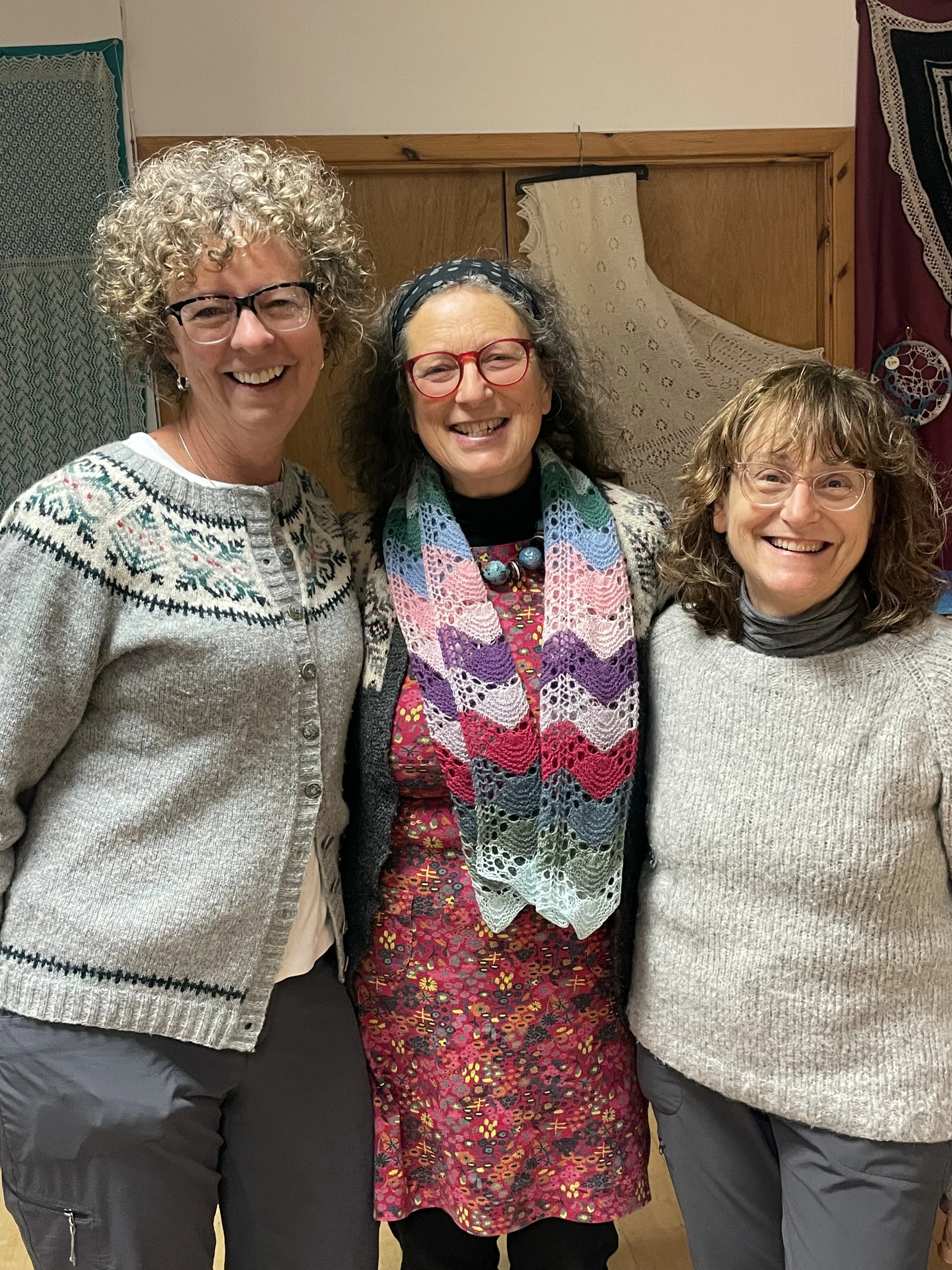 Three smiling middle-aged women standing together, wearing glasses and sweaters, in front of decorative textiles.