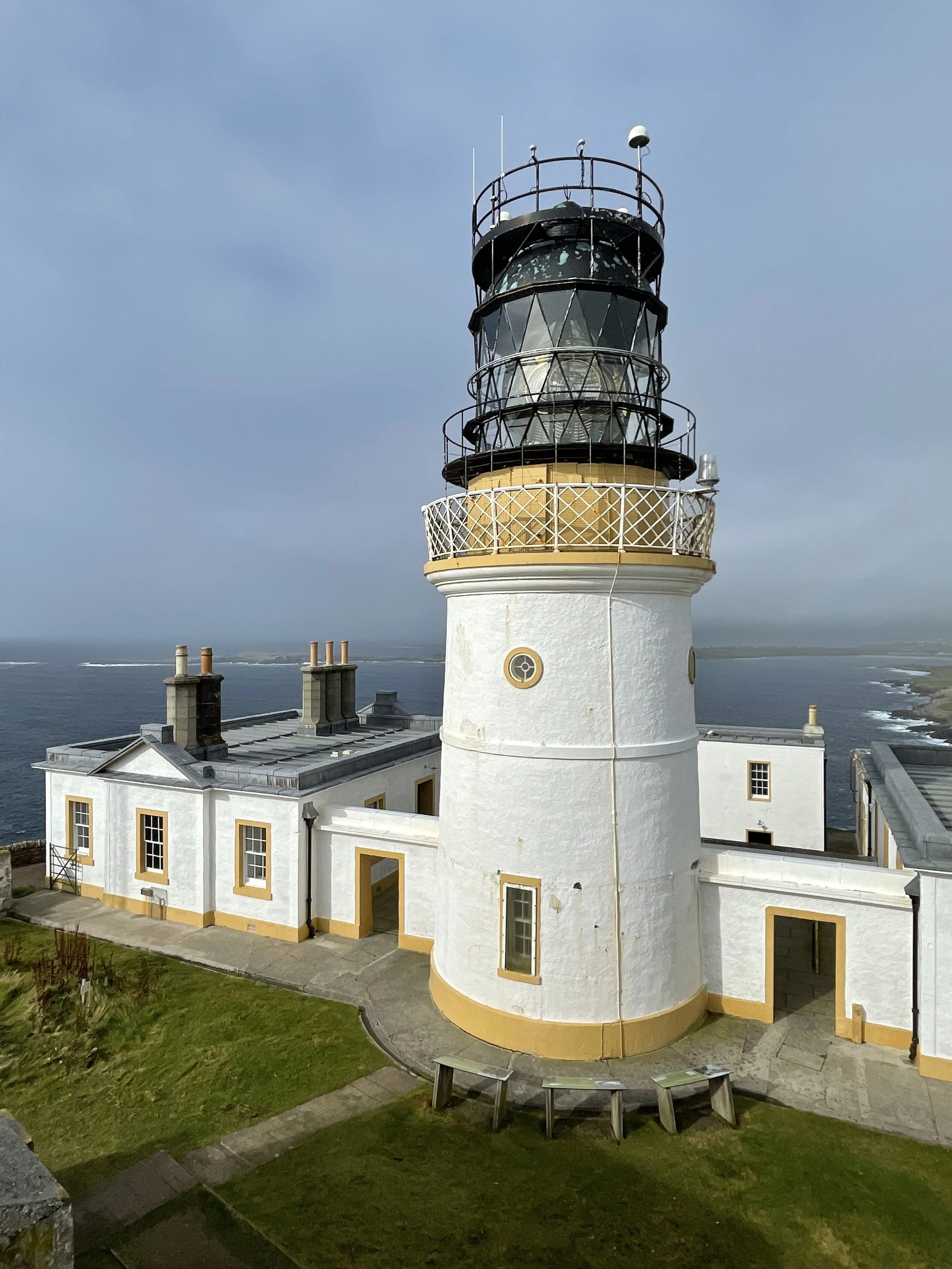 White lighthouse with yellow accents near the coast, surrounded by additional buildings, grassy area, and ocean view.