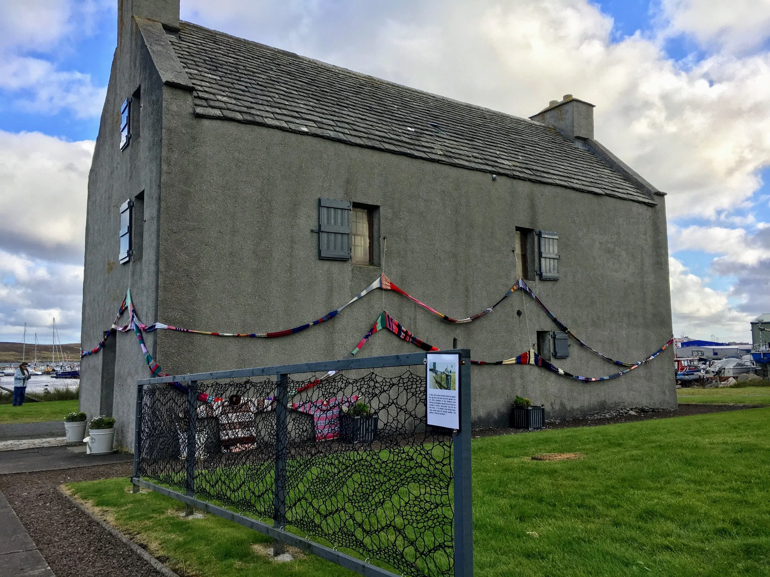 Gray stone house with colorful fabric decorations, fenced yard, and cloudy sky in background.