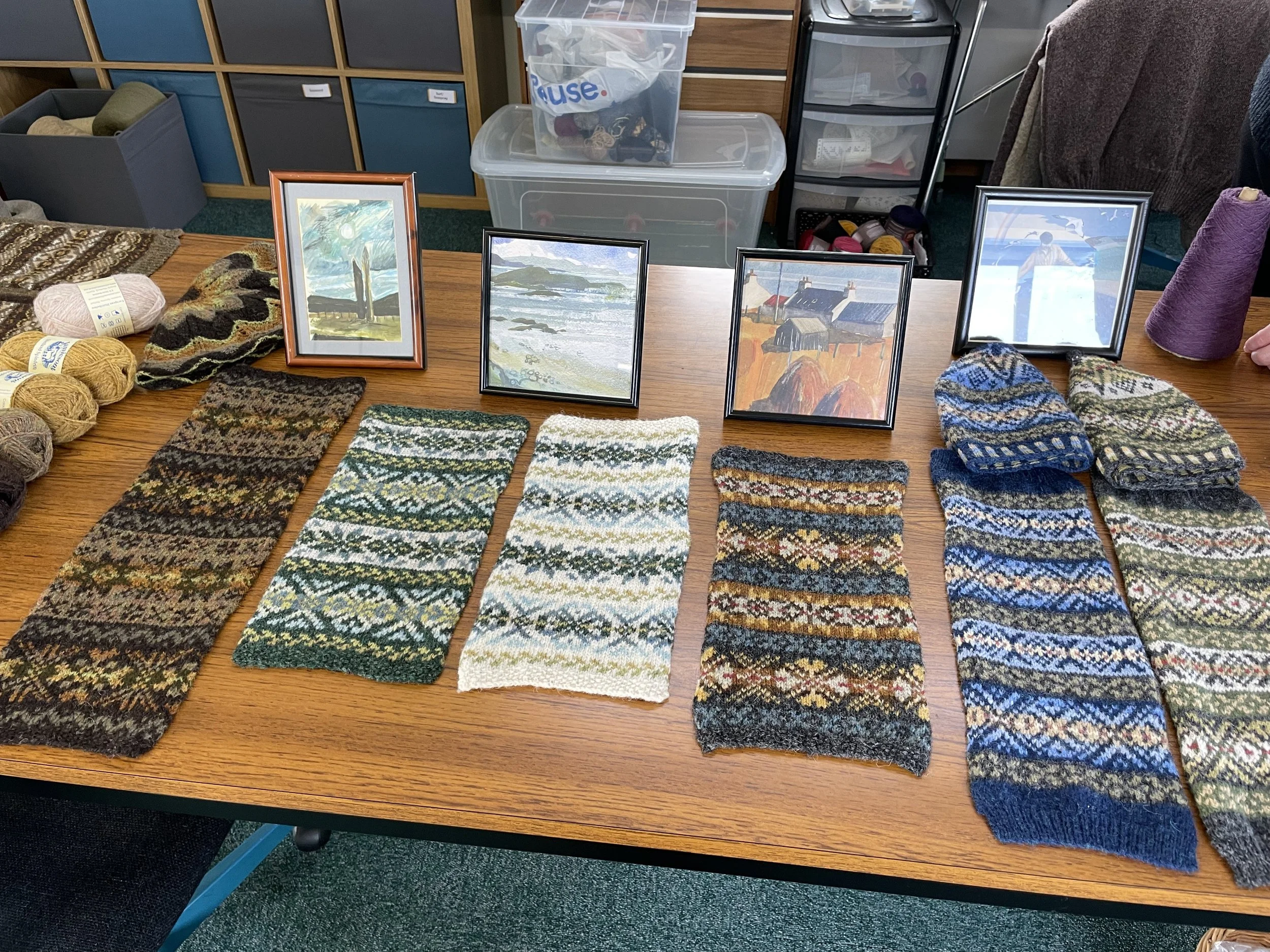 A tabletop display showing knitted fabric swatches and knitted hats with various intricate patterns. Yarn balls and art pieces are also present on the table. Shelves in the background hold containers and textile materials.