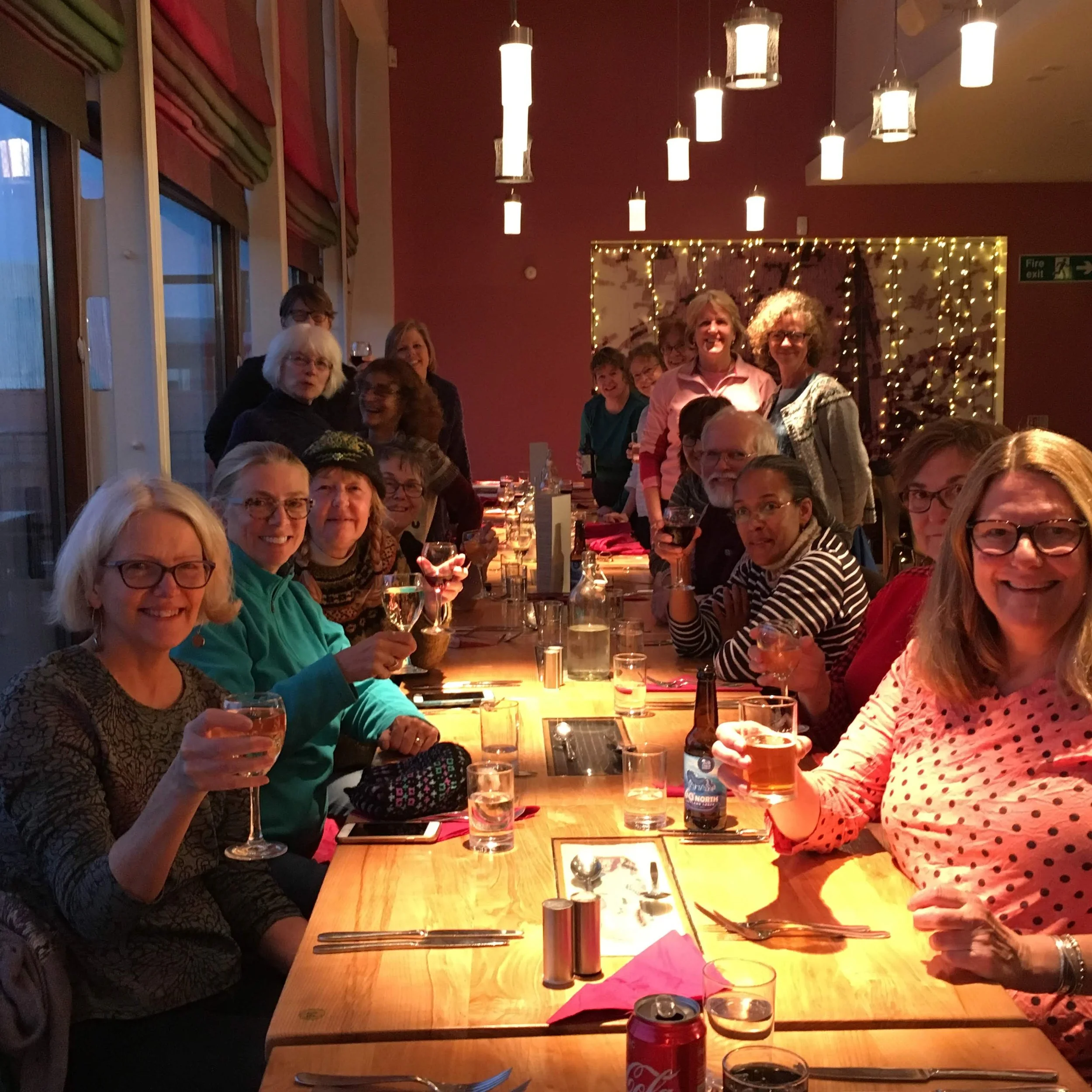 A group of people sitting at a long dining table, holding glasses and smiling, in a warmly lit restaurant setting.