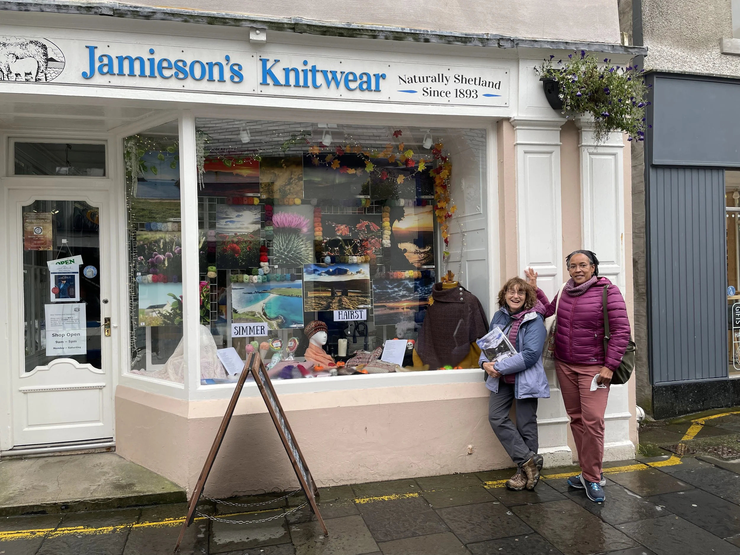 Two people standing outside Jamieson's Knitwear shop in Shetland. The shop window displays colorful wool and knitwear items, with scenic pictures on the wall. There's a small street sign and hanging flower basket.