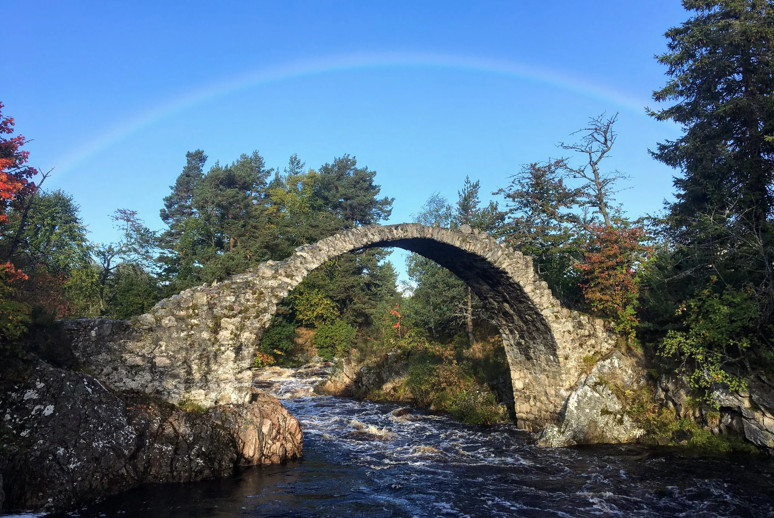 Stone arch bridge over a river with a rainbow in the background, surrounded by trees.