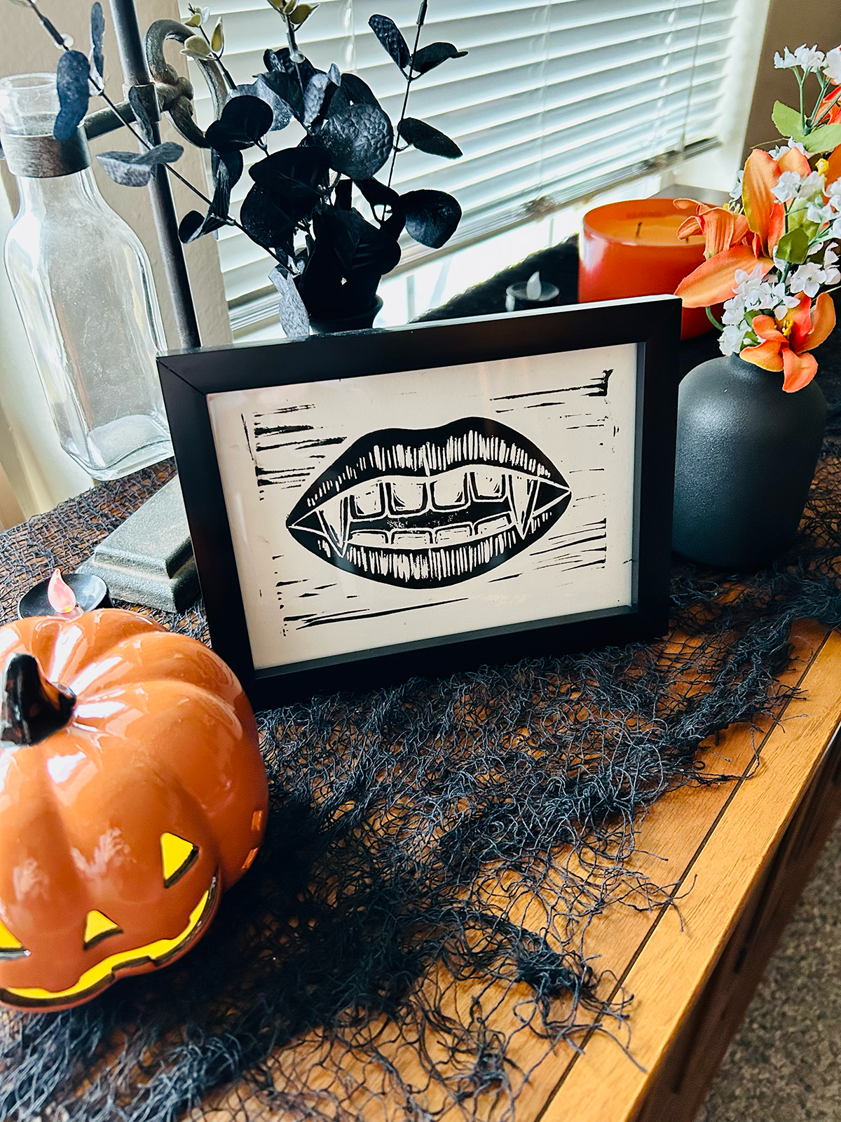 Decorative Halloween-themed display on a wooden table with a black lace table runner. It includes a black framed sign with a stylized depiction of vampire teeth, an orange pumpkin with a carved face, a black vase with orange and white flowers, an ora
